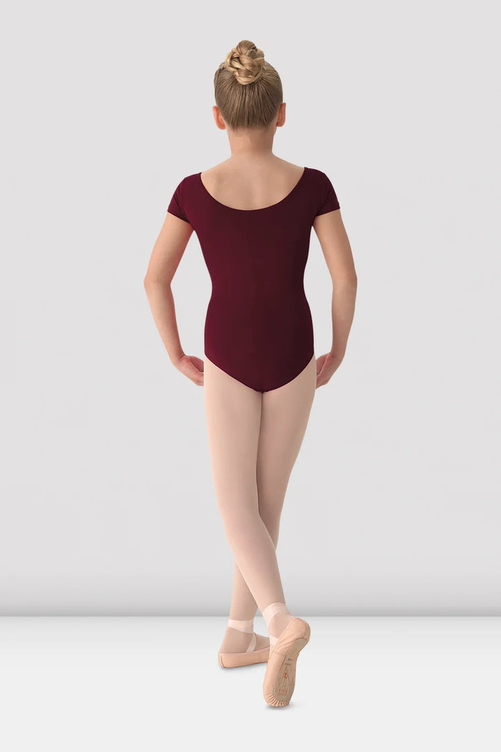 A young girl dancer shown from the back, wearing a short-sleeved burgundy leotard, beige tights, and pink pointe shoes against a light gray studio background.