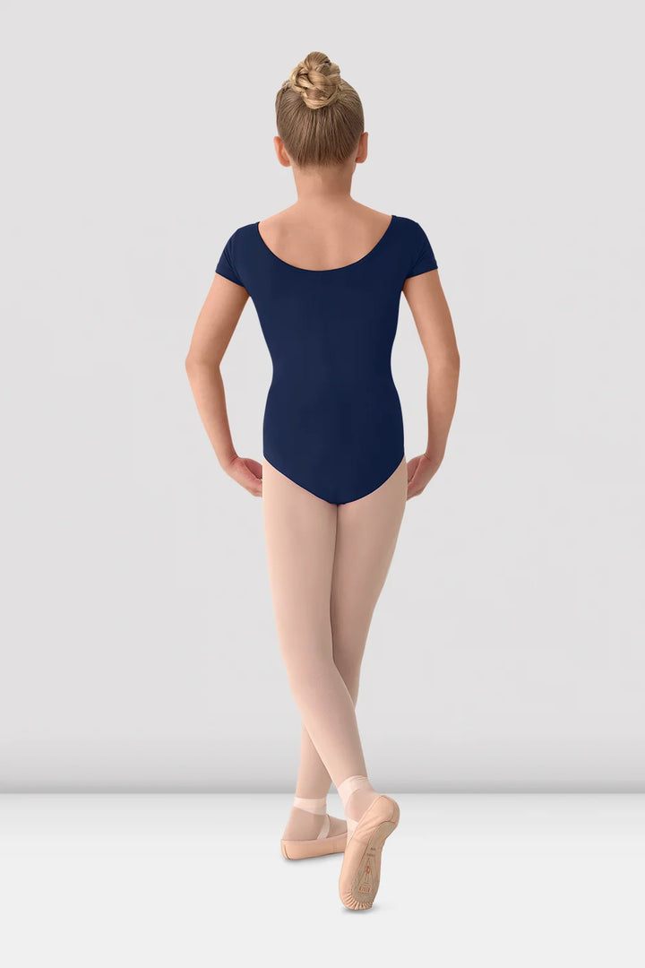 A young ballerina viewed from behind, wearing a navy blue short-sleeve leotard, nude tights, and pink pointe shoes against a light gray studio background.
