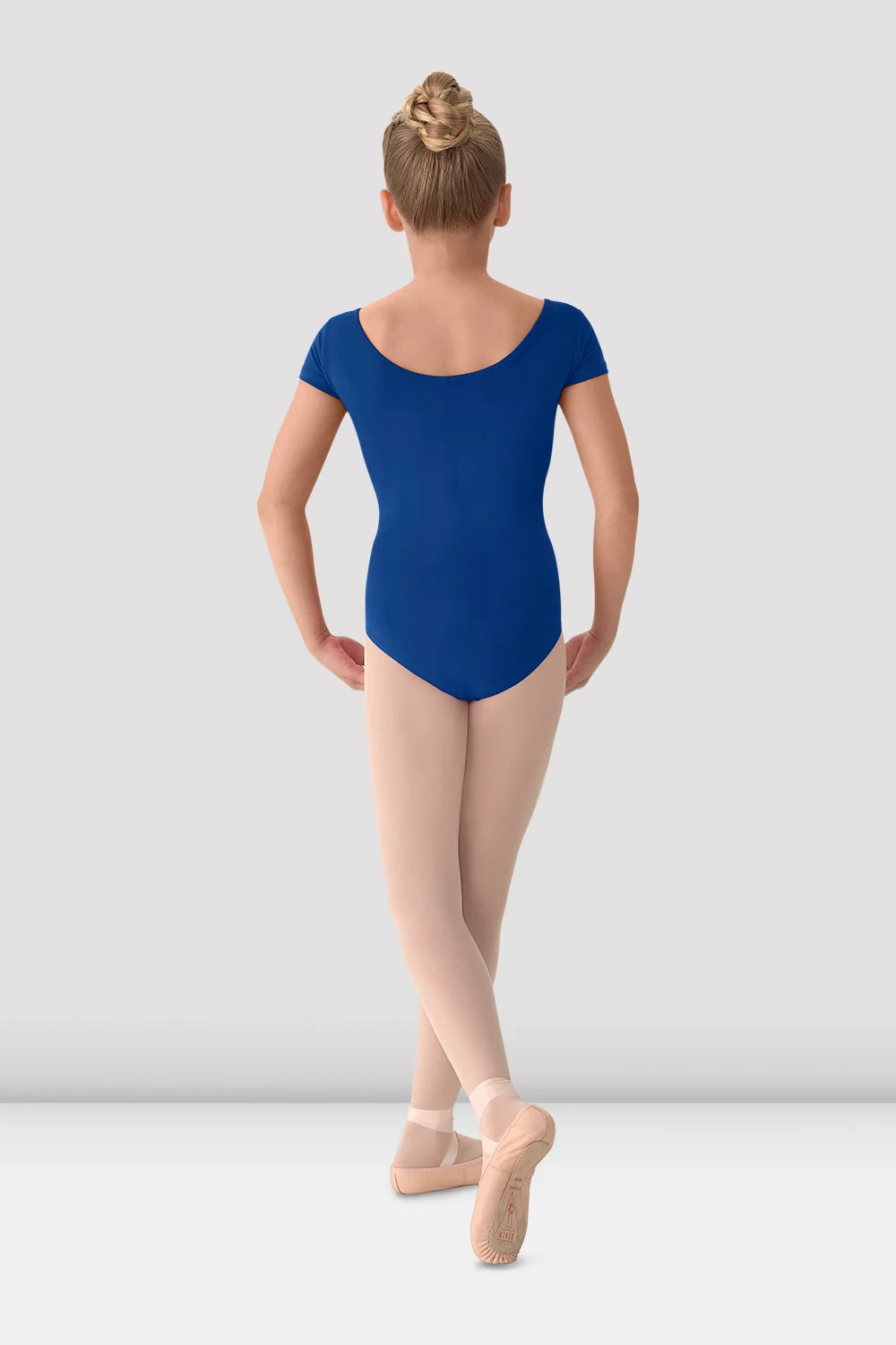 Young girl in a blue leotard and beige tights standing against a white background