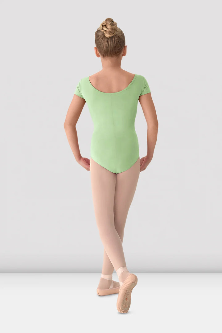 A young girl dancer viewed from behind, wearing a light green short-sleeve leotard, beige tights, and pink pointe shoes against a light gray studio background.