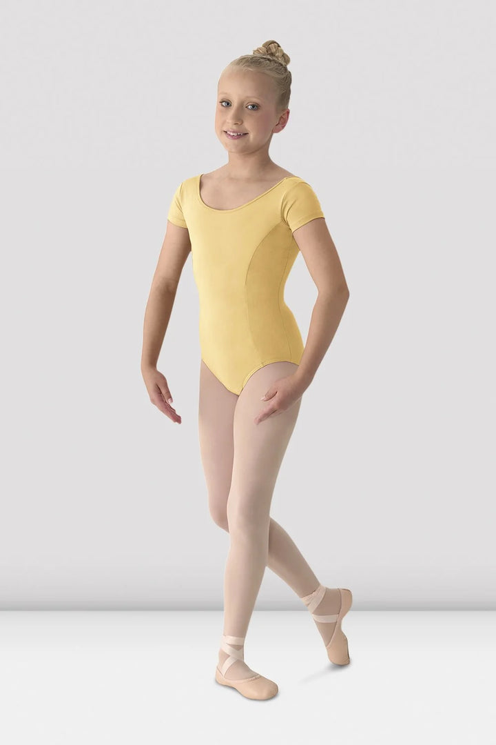 A young girl dancer wearing a pale yellow short-sleeve leotard, nude tights, and pointe shoes, posing against a light gray studio background.