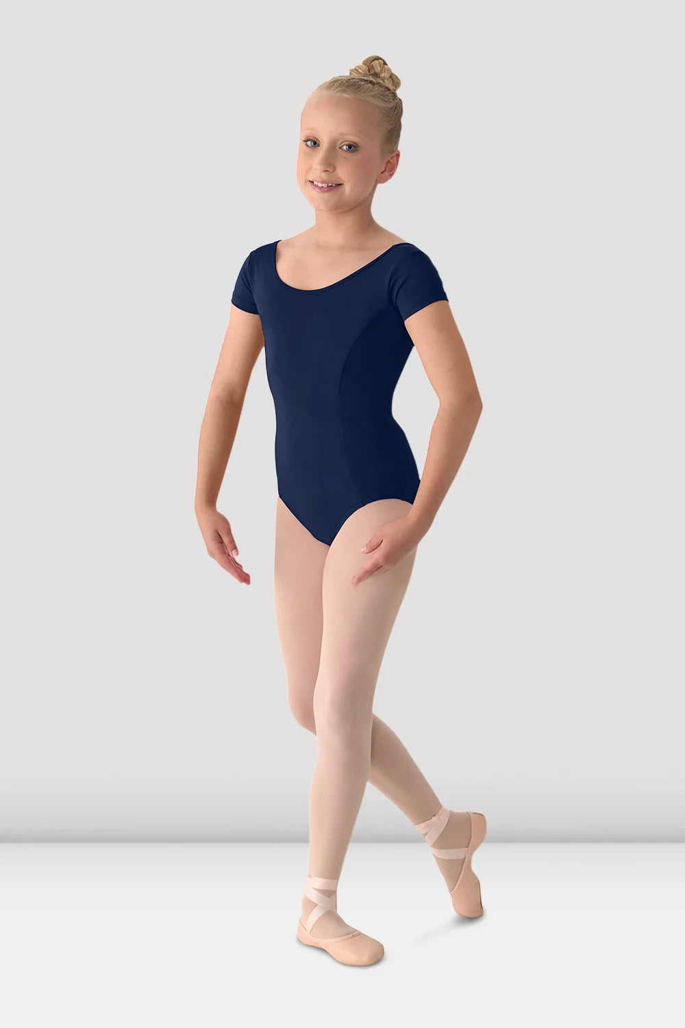 A young girl dancer wearing a navy blue short-sleeve leotard, pink tights, and pink pointe shoes, posing against a light gray studio background.