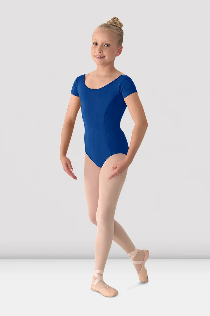 A young girl dancer wearing a royal blue short-sleeve leotard, nude tights, and pink pointe shoes, posing against a light gray studio background.