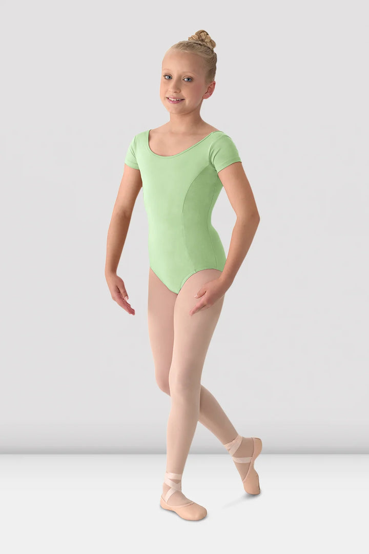 A young girl dancer wearing a light green short-sleeve leotard, pink tights, and pink pointe shoes, posing against a light gray studio background.