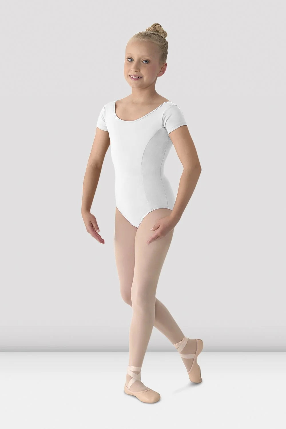 A young girl dancer wearing a white short-sleeve leotard, nude tights, and pink pointe shoes, posing against a light gray studio background.