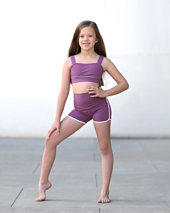 Young girl in a purple sports bra and shorts posing on a concrete floor.