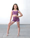 Young girl in a purple sports bra and shorts posing on a concrete floor.
