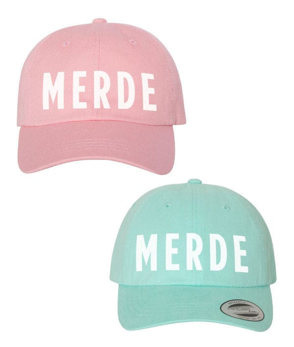 Pink and teal baseball caps with 'MERDE' text on a white background