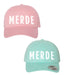 Pink and teal baseball caps with 'MERDE' text on a white background