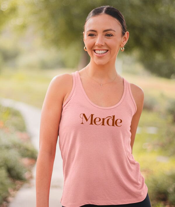 Woman wearing a pink tank top with 'Merde' printed on it, standing outdoors.