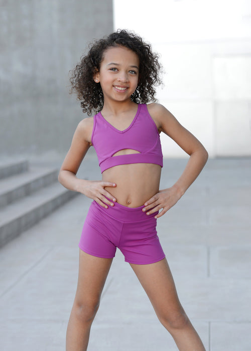 Young girl wearing a purple sports bra and shorts set standing outdoors.
