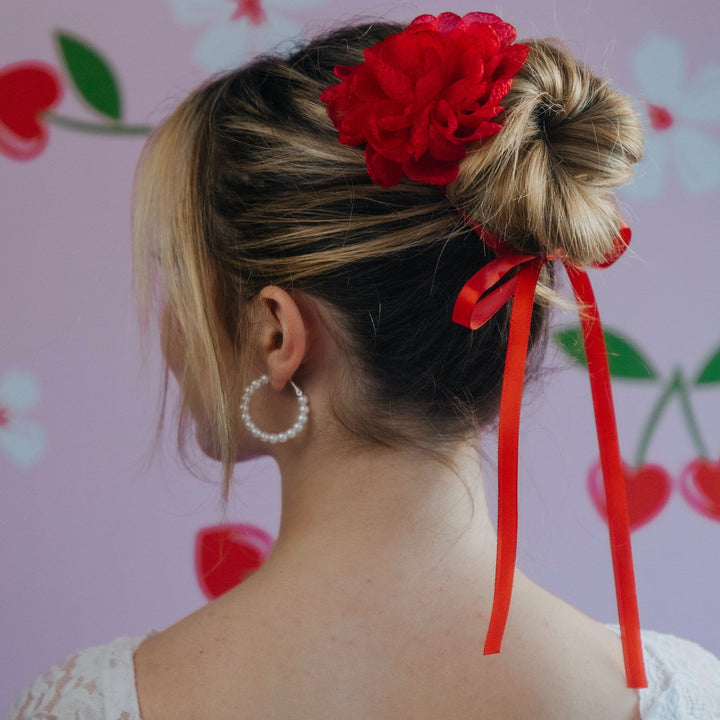 Back view of a person with a styled hair bun featuring a red flower and ribbons against a floral background.