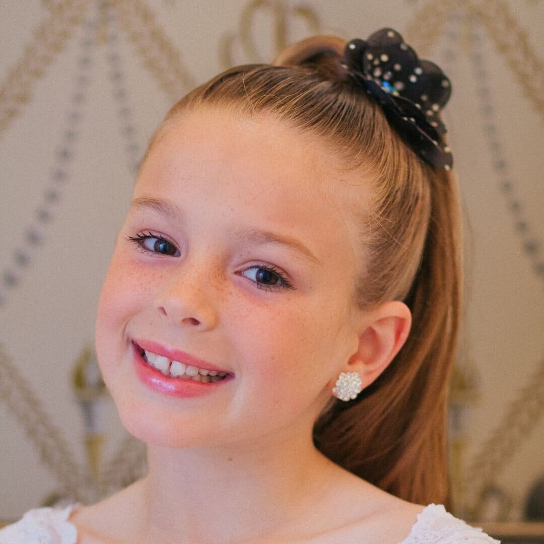 Young girl with a ponytail and decorative hair bow, wearing earrings, against a decorative wall.