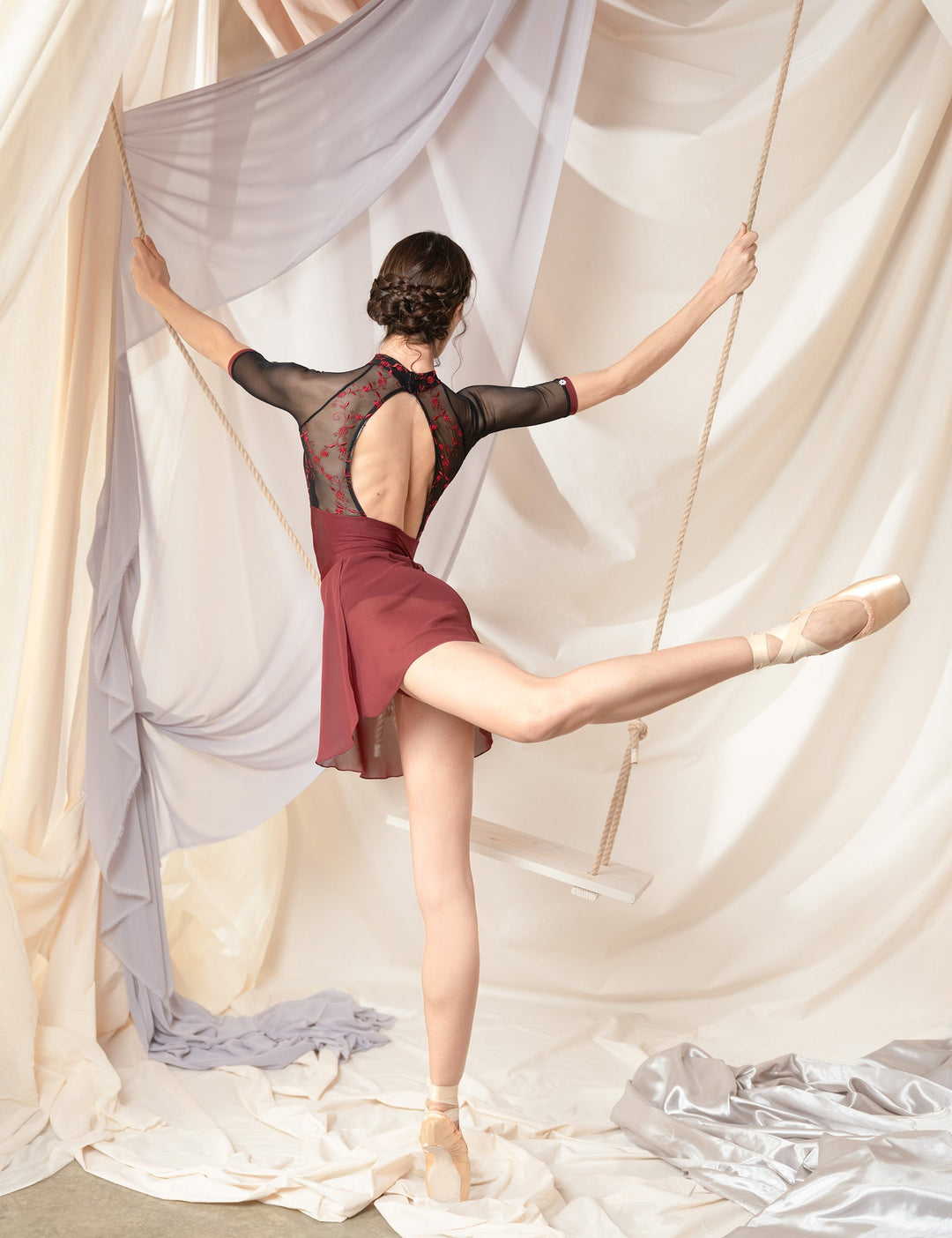 Ballerina in a red dress with sheer overlay posing against draped fabric.