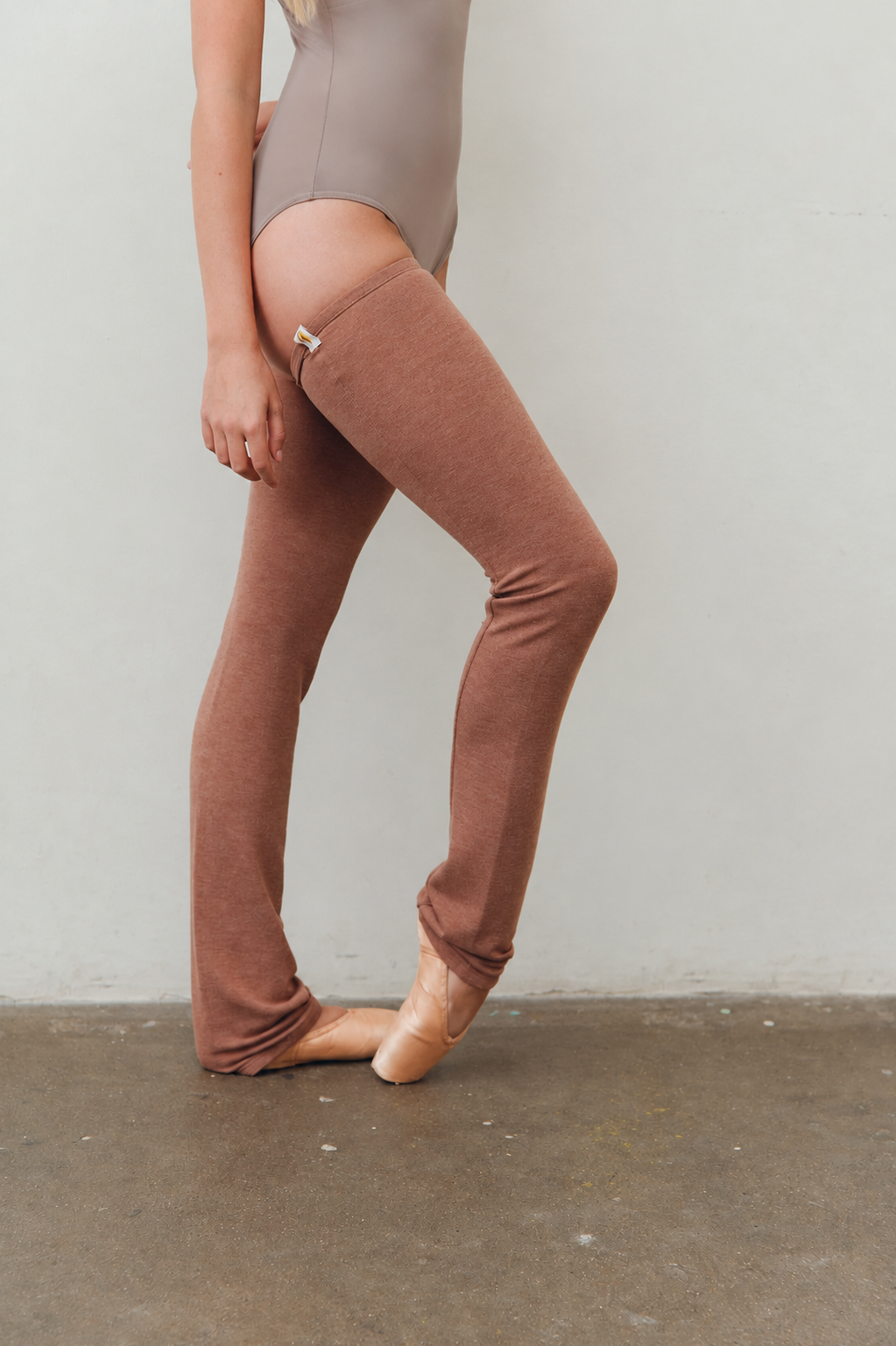 A model wearing dusty rose tights with a small gold zipper detail at the top, paired with a taupe bodysuit and ballet slippers, standing on a concrete floor against a light gray wall.