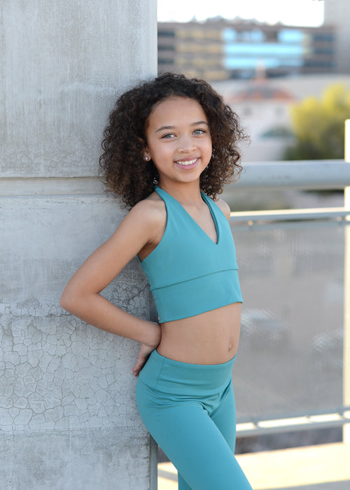 Young girl in teal athletic wear leaning against a stone column with a blurred cityscape background