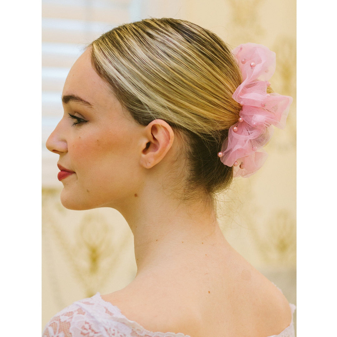Woman with blonde hair tied back with a pink scrunchie against a blurred natural background