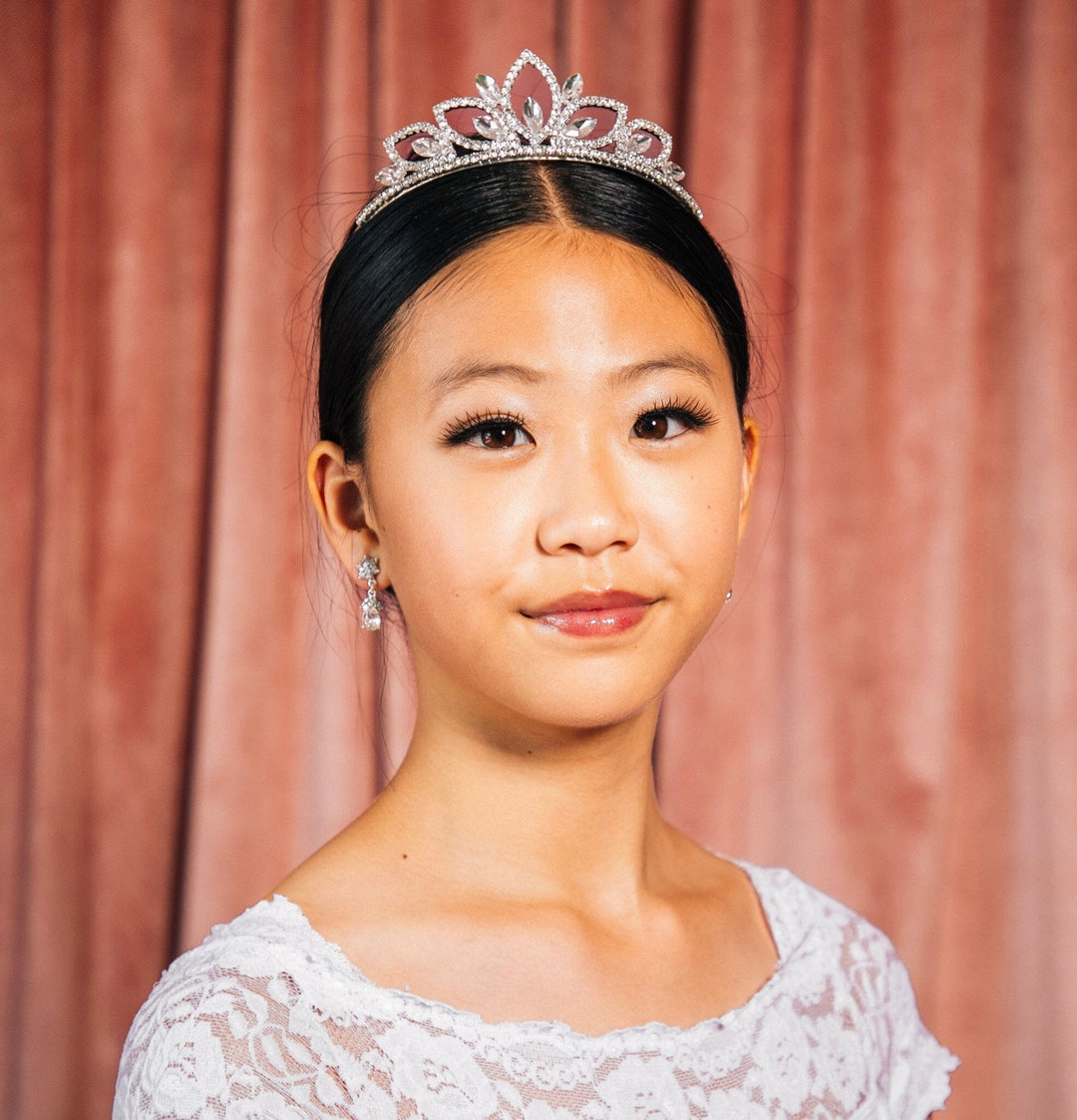 Young woman wearing a tiara and white lace top against a pink curtain background