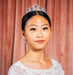 Young woman wearing a tiara and white lace top against a pink curtain background