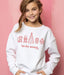 Young girl wearing a white sweatshirt with red Christmas-themed graphics and text on a pink background