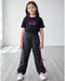 Young girl wearing a black t-shirt with 'Purple Pixies' text and black pants with pink accents on a light wooden floor.