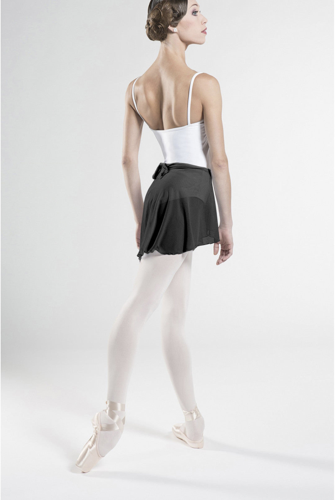 A female ballet dancer shown from the back, wearing a white camisole, a black tutu skirt, white tights, and pointe shoes against a plain light gray studio background.