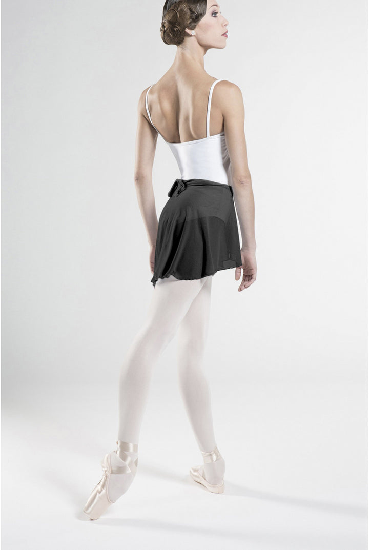 A female ballet dancer shown from the back, wearing a white camisole, a black tutu skirt, white tights, and pointe shoes against a plain light gray studio background.