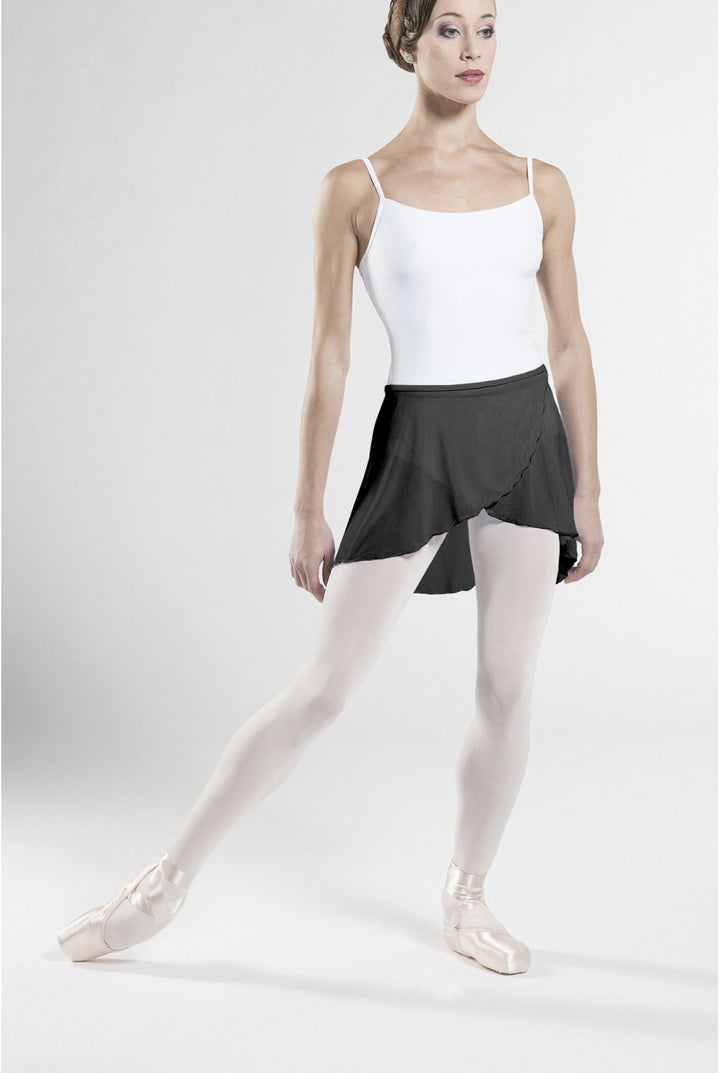 A female dancer posing in a white camisole, a black ruffled ballet skirt, white tights, and pointe shoes against a light gray studio background.