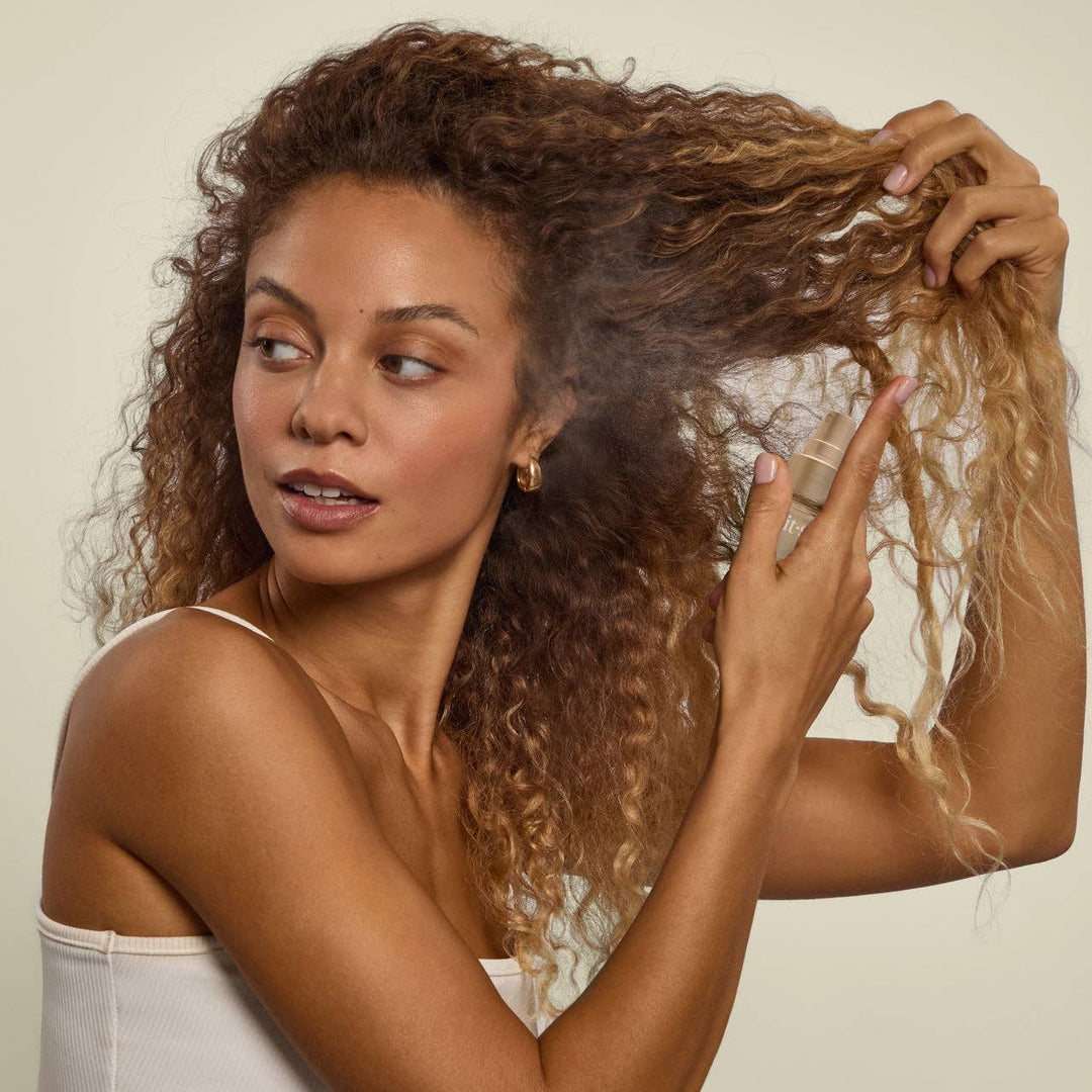 Woman with curly hair holding a bottle of hair product against a neutral background