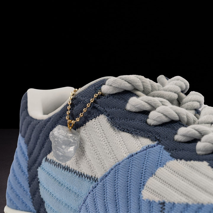 Close-up of a blue and white sneaker with braided laces and a gold chain detail on a black background.
