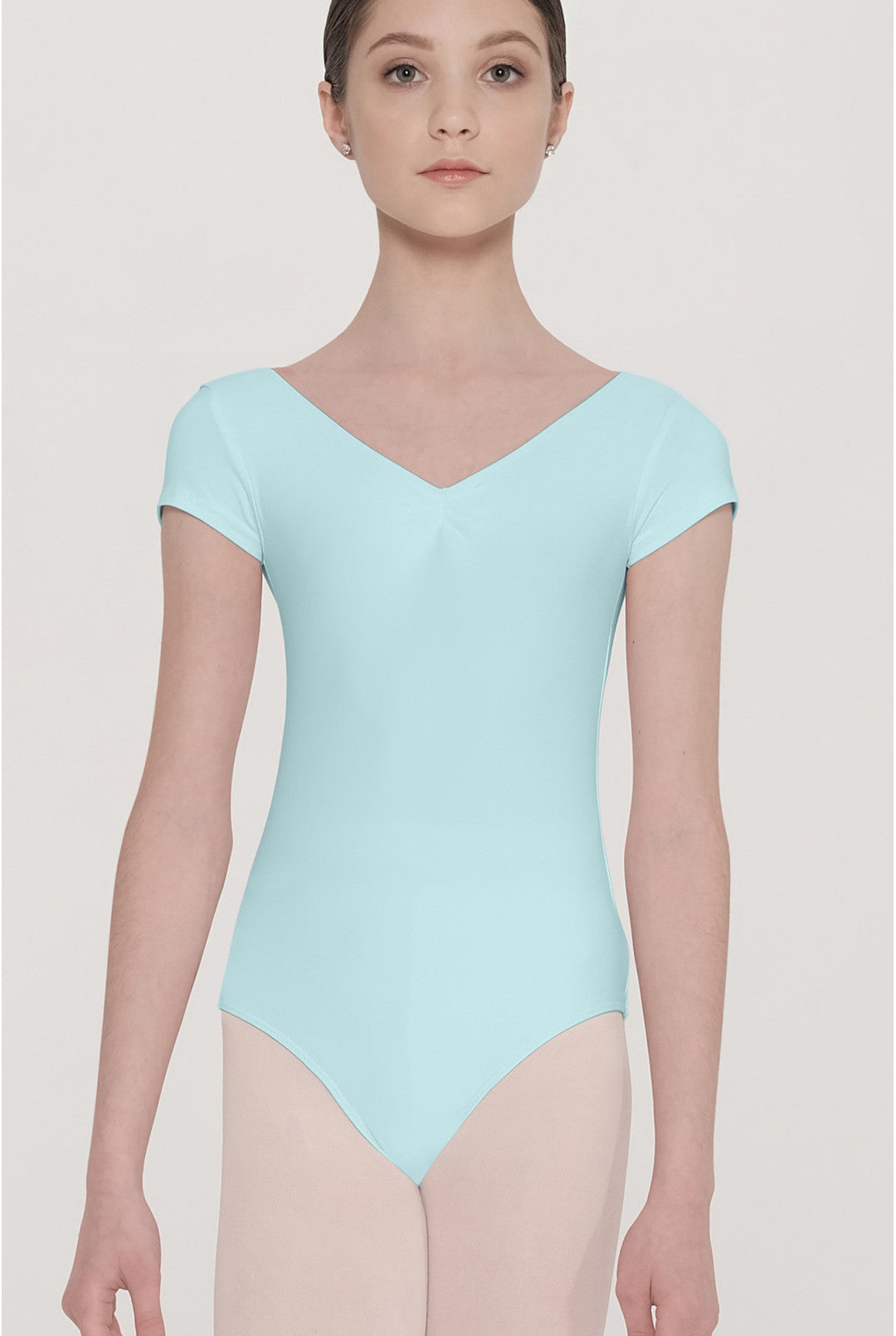 A female dancer wearing a light mint blue short-sleeve leotard and nude tights, standing against a solid light beige background.