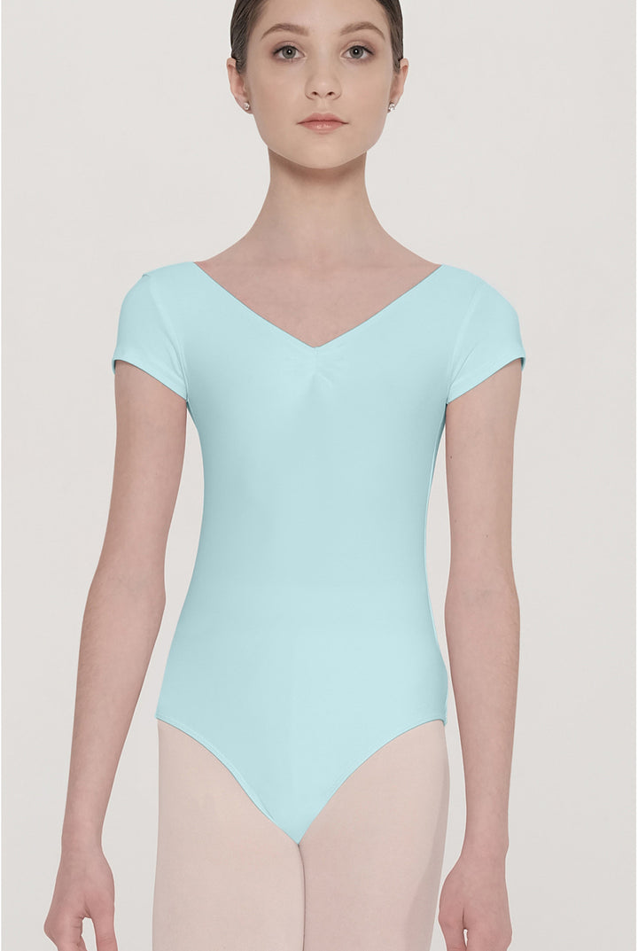 A female dancer wearing a light mint blue short-sleeve leotard and nude tights, standing against a solid light beige background.