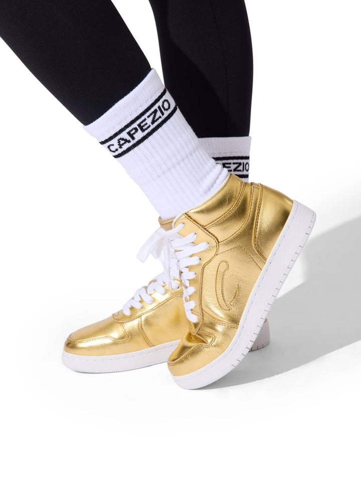 Gold high-top sneakers with white soles worn with black leggings and white socks featuring 'Capezio' branding.