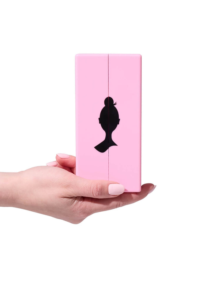 Pink makeup compact with black silhouette design held by a hand on a white background