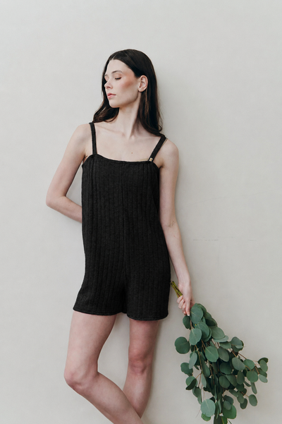 A woman wearing a black ribbed knit camisole romper with thin shoulder straps, leaning against a light beige wall and holding a green eucalyptus branch.