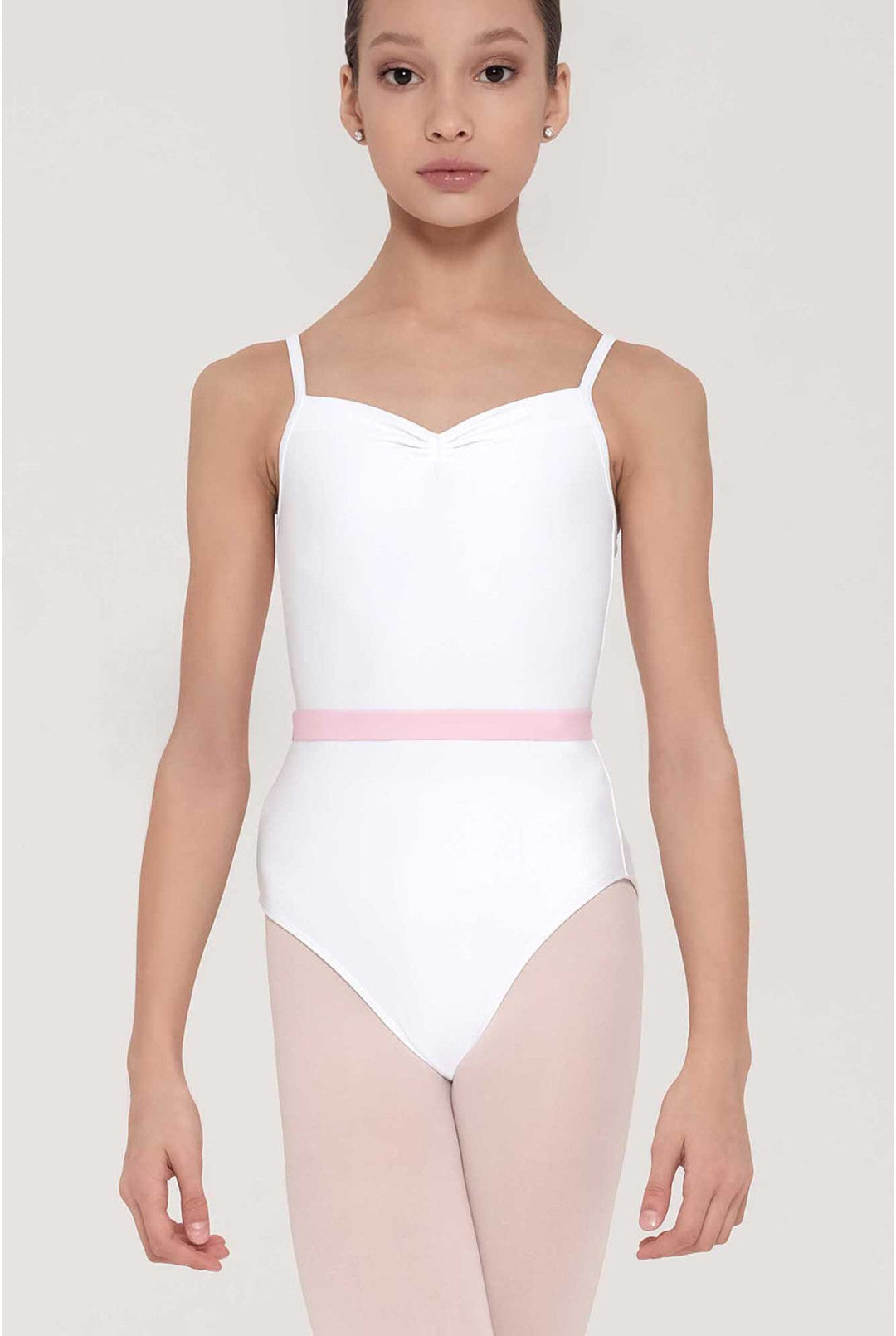 A young girl modeling a white ballet leotard with thin shoulder straps and a light pink waistband, paired with pale pink tights, against a light gray studio background.