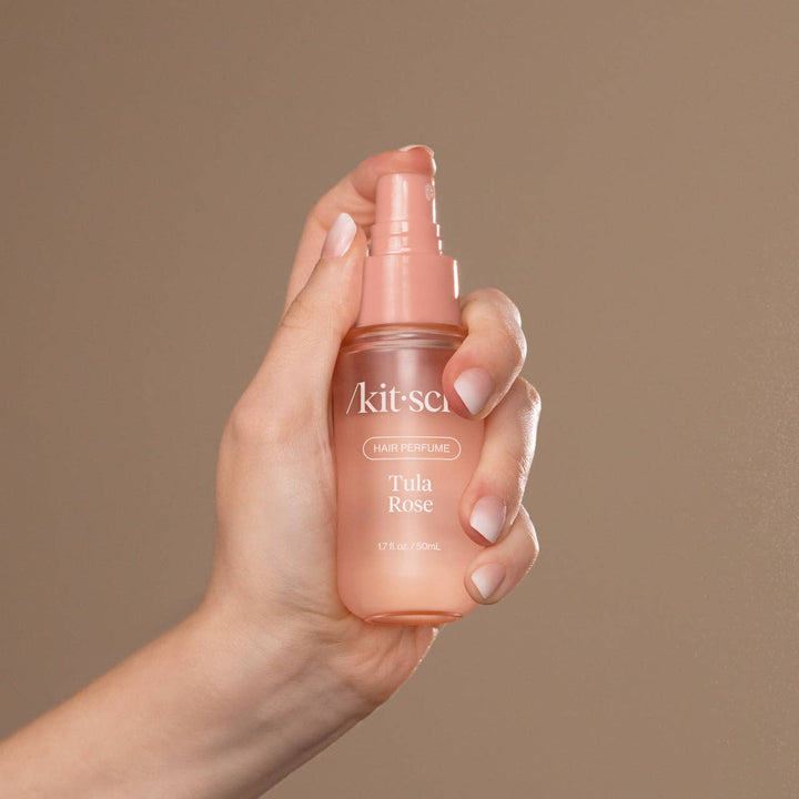 Hand holding a pink bottle of 'Akitsuki' Tula Rose hair perfume against a brown background