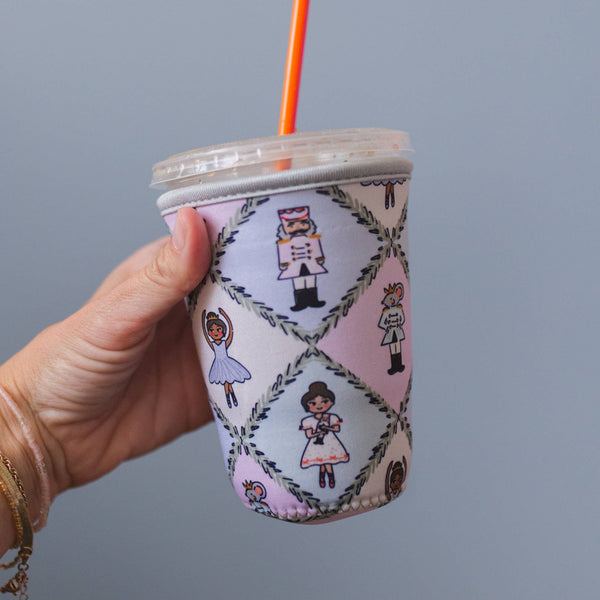 A hand holding a clear plastic cup with a clear lid and an orange straw, wrapped in a pastel quilted fabric sleeve featuring cartoon nurse and nursemaid illustrations on a light gray background.