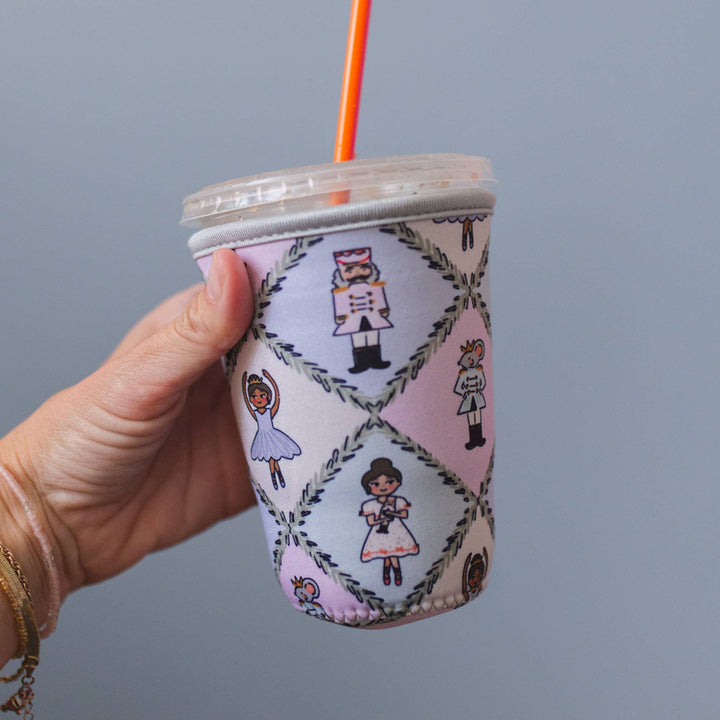 A hand holding a clear plastic cup with a clear lid and an orange straw, wrapped in a pastel quilted fabric sleeve featuring cartoon nurse and nursemaid illustrations on a light gray background.