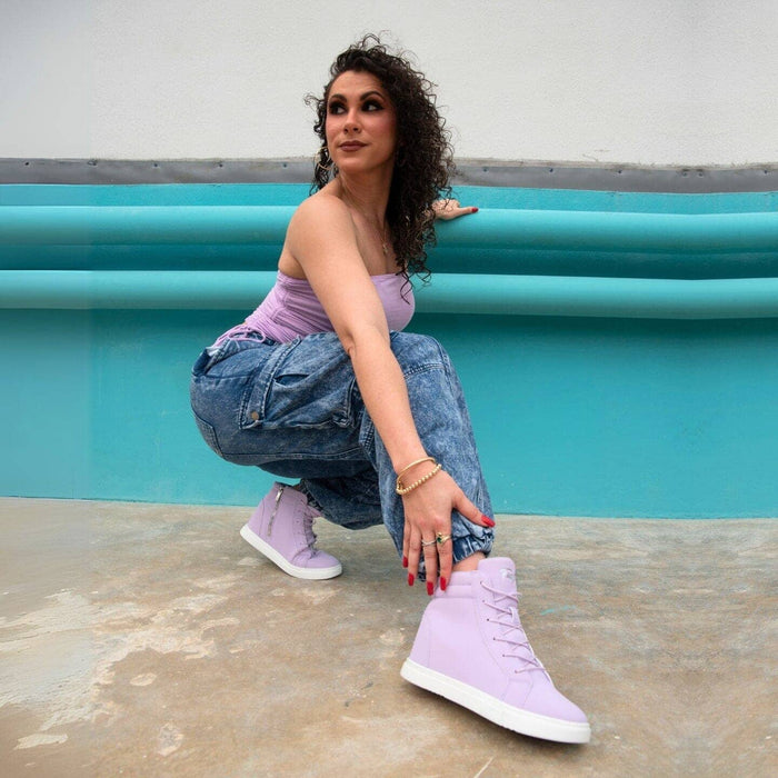 Woman wearing lavender high-top sneakers and a matching top, sitting against a turquoise wall.