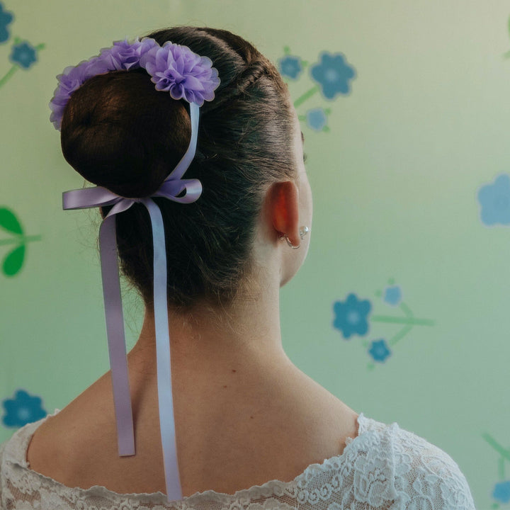 Person with a bun hairstyle featuring purple flowers and ribbons against a floral-patterned wall.